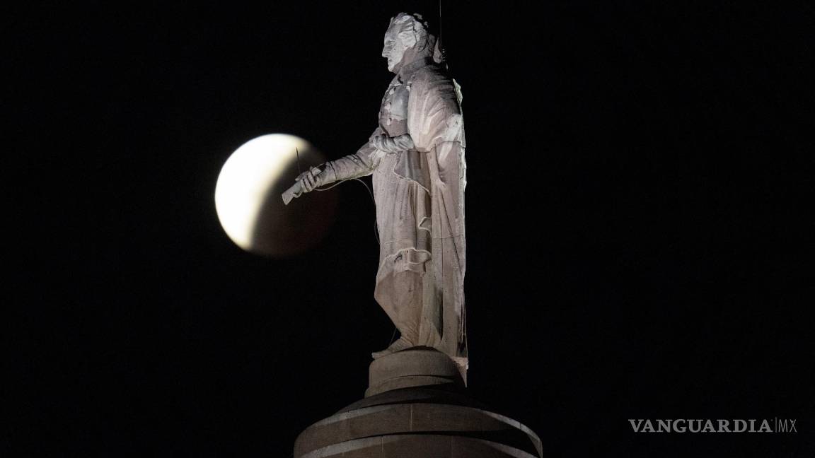 $!La sombra de la tierra cubre la luna llena durante un eclipse lunar parcial visible cerca de una estatua de George Washington en lo alto del Monumento a Washington de Baltimore en Baltimore. AP/Julio Cortez