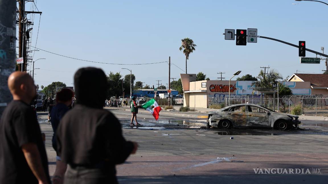 $!Un manifestante scon una bandera mexicana junto a un automóvil quemado durante un enfrentamiento entre manifestantes y agentes federales en Paramount.