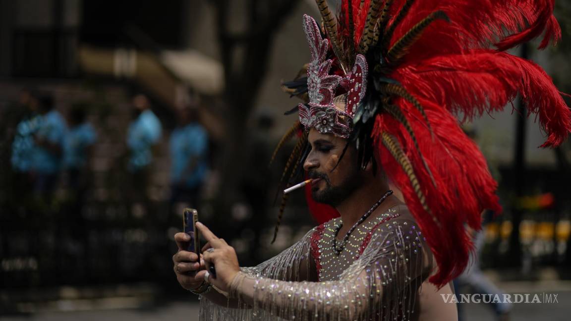 $!Un participante, con un tocado azteca y fumando un cigarrillo, posa para una selfie mientras participa en la marcha anual del Orgullo en la Ciudad de México.