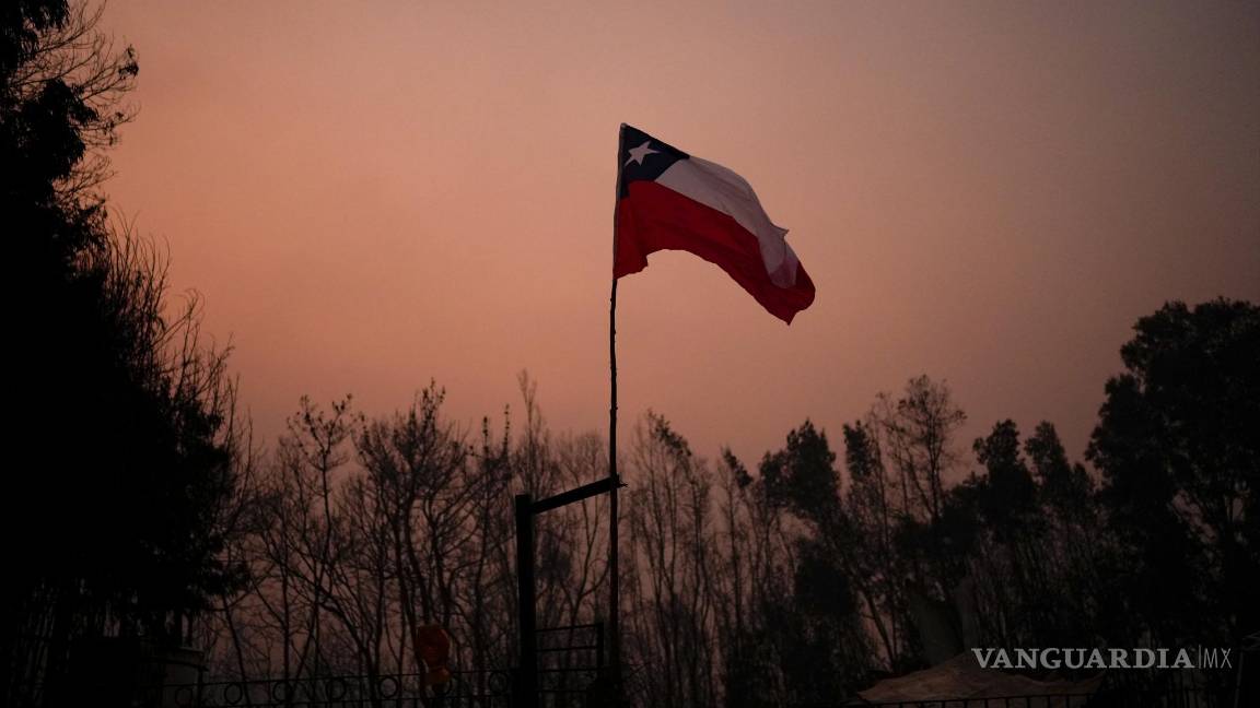 $!Una bandera chilena colocada en medio de árboles carbonizados bajo un cielo rojo causado por incendios forestales en Santa Juana, Chile.