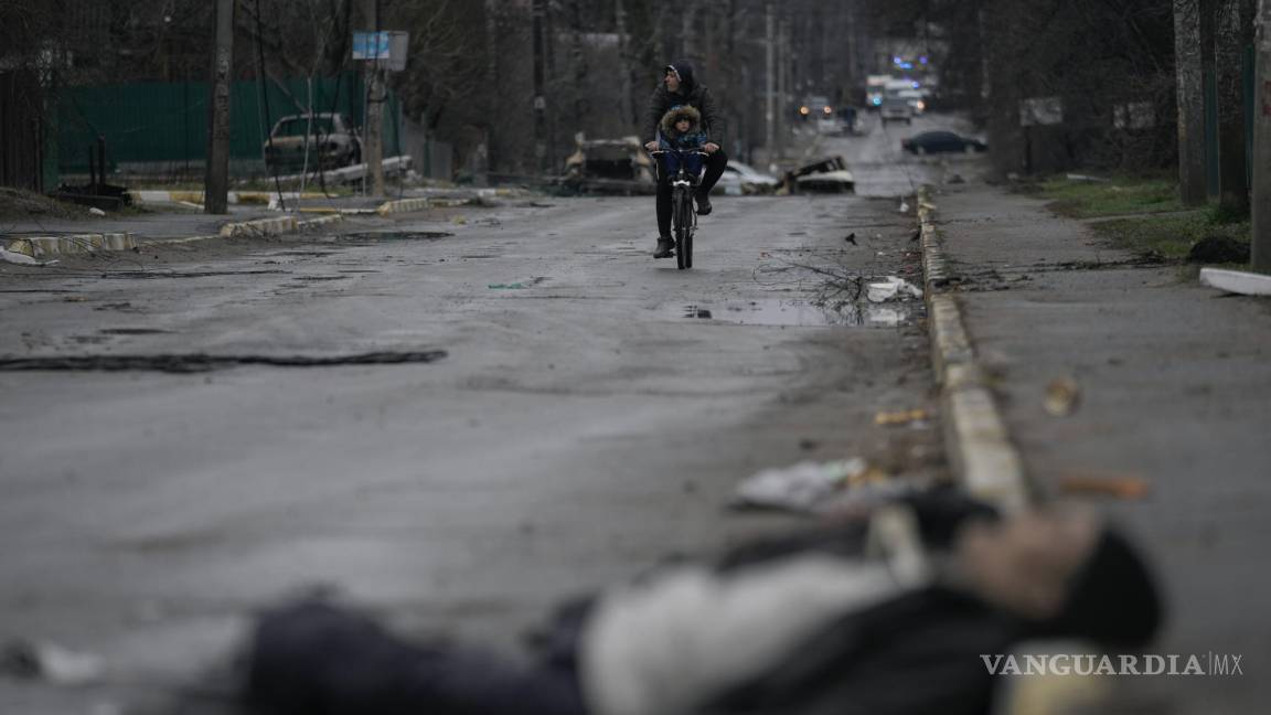 $!Un hombre y un niño montan en bicicleta por una calle donde se ven cuerpos de civiles en Bucha que estuvo ocupado por fuerzas rusas, en Ucrania.