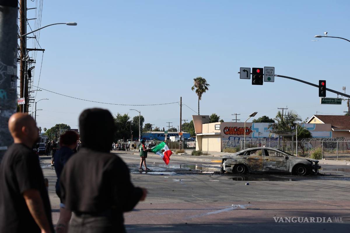 $!Un manifestante scon una bandera mexicana junto a un automóvil quemado durante un enfrentamiento entre manifestantes y agentes federales en Paramount.