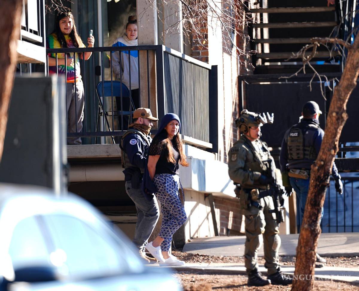 $!Mientras un vecino registra el momento con su teléfono, una mujer es sacada por autoridades de un departamento durante una redada en el este de Denver.