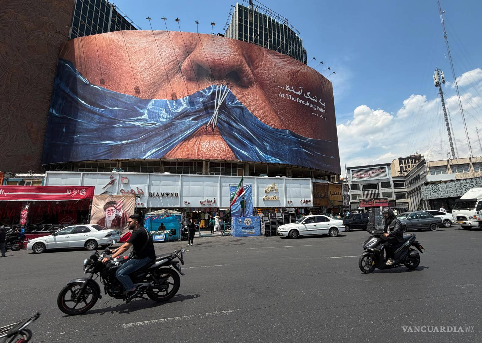 $!Vehículos circulan junto a un mural publicitario con una imagen del estrecho de Ormuz y los labios cosidos del presidente Donald Trump, en Teherán.