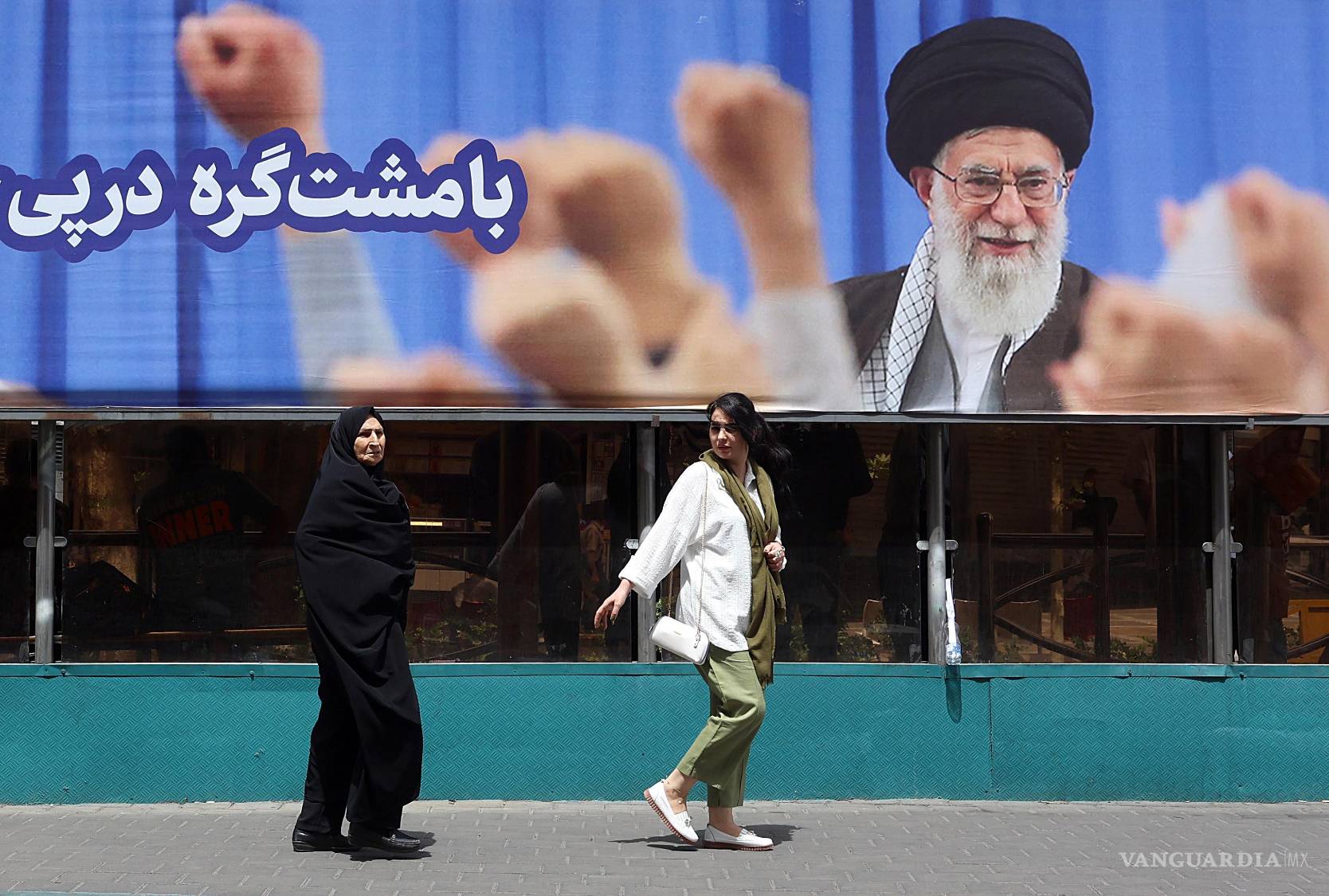 $!Mujeres iraníes pasan junto a un gran cartel del difunto líder supremo iraní, el ayatolá Ali Jamenei, antes de su funeral en Teherán, Irán, el 2 de julio de 2026.
