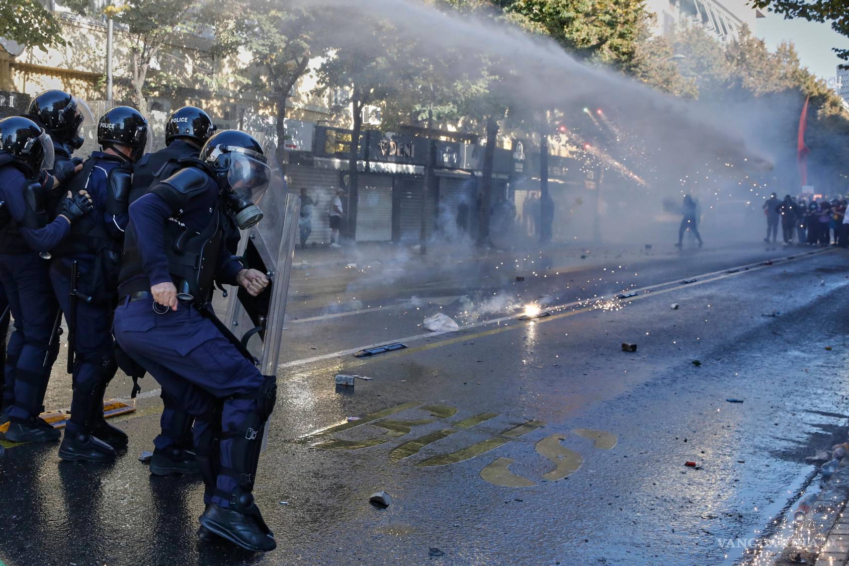 $!Agentes antidisturbios se resguardan tras sus escudos mientras estallan fuegos artificiales cerca y un cañón de agua rocía a los manifestantes durante una protesta antigubernamental en Tirana, Albania, el martes 20 de octubre de 2026.