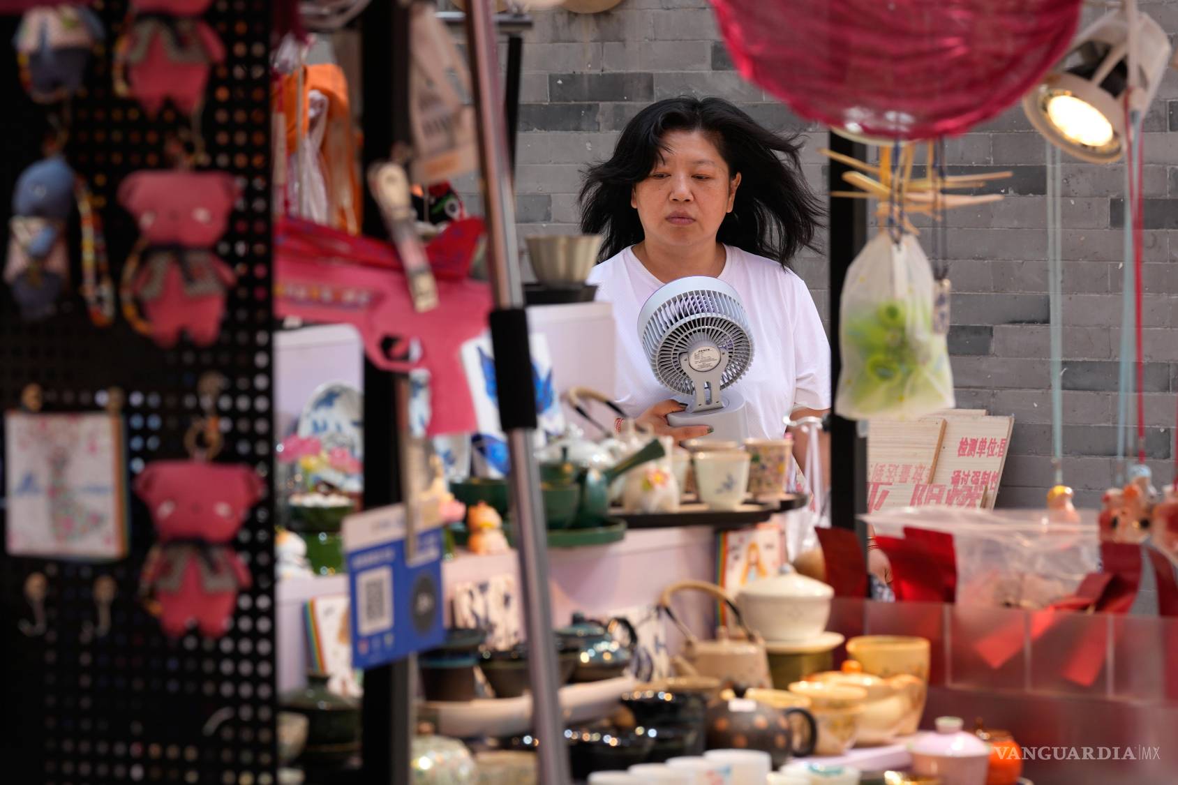 $!Una vendedora se refresca con un ventilador eléctrico portátil mientras espera clientes en su tienda de recuerdos en Pekín, China.