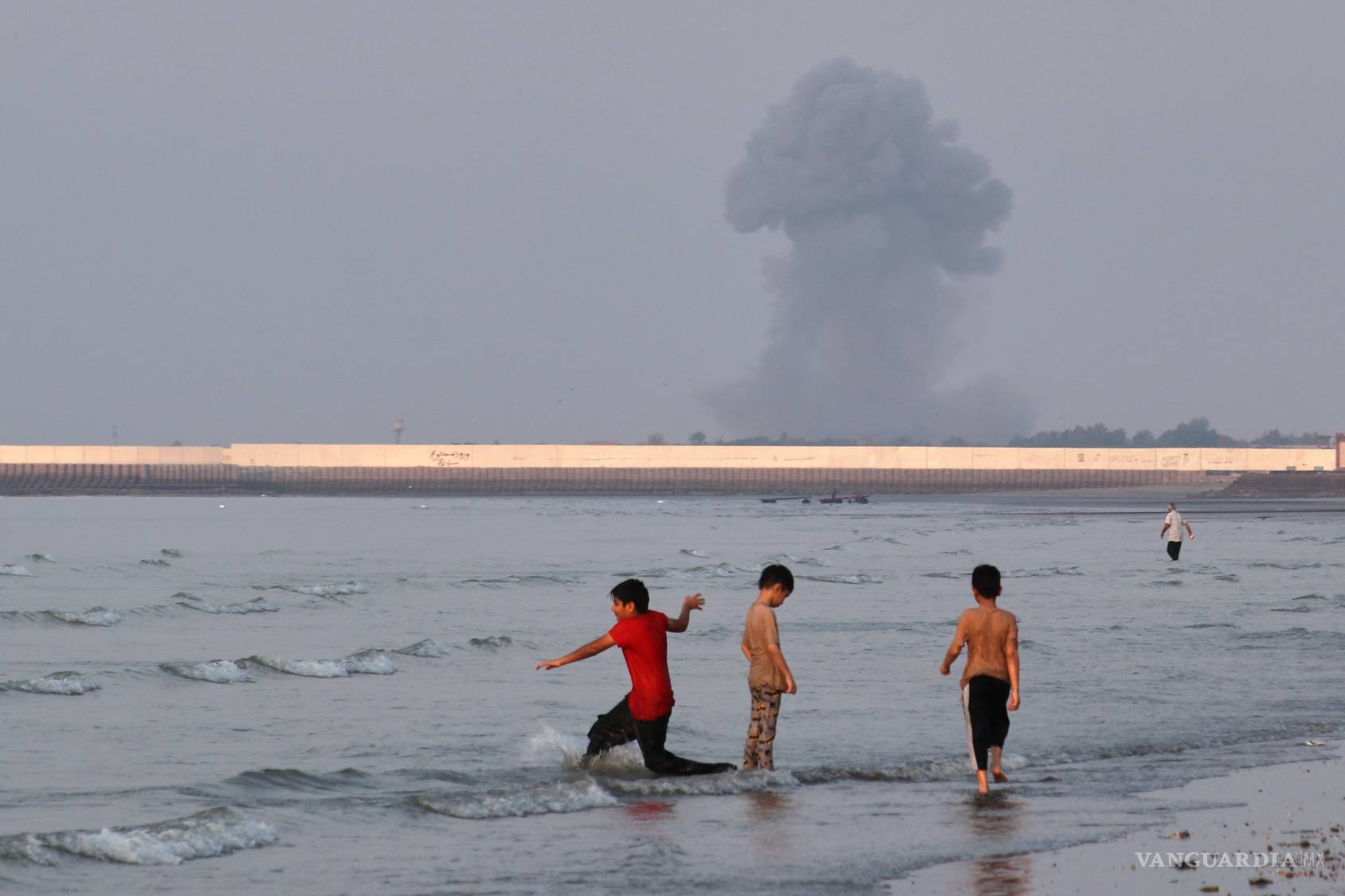 $!Tres niños juegan en el agua en el estrecho de Ormuz mientras al fondo se alza una columna de humo de una explosión, cerca de Bandar Abbas, Irán.