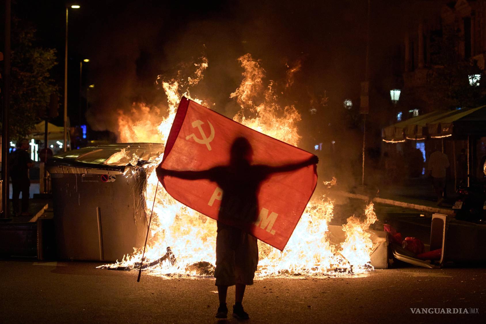 $!Un manifestante sostiene una bandera junto a contenedores de basura en llamas durante enfrentamientos con la policía tras una manifestación por la crisis de vivienda en España.