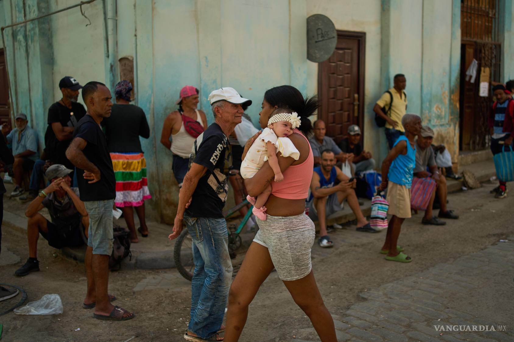 $!Una mujer camina con un bebé en brazos junto a personas que hacen fila para comprar pan durante un apagón en La Habana, Cuba.