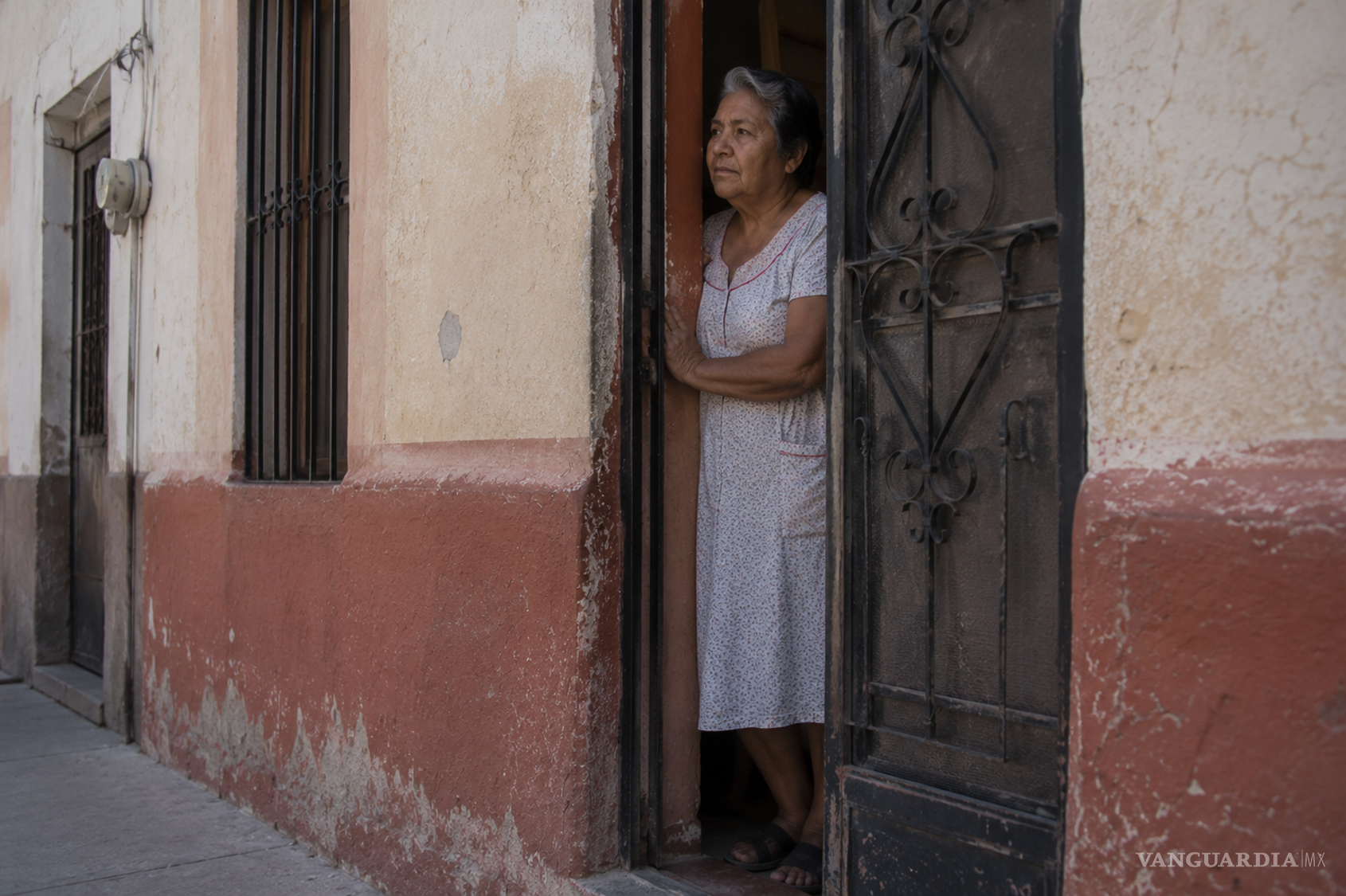 $!Una mujer observa la calle desde la penumbra de su casa. La puerta permanece abierta, pero el umbral conserva su función de límite entre la intimidad doméstica y la vida pública del barrio. Ilustración generada con IA.