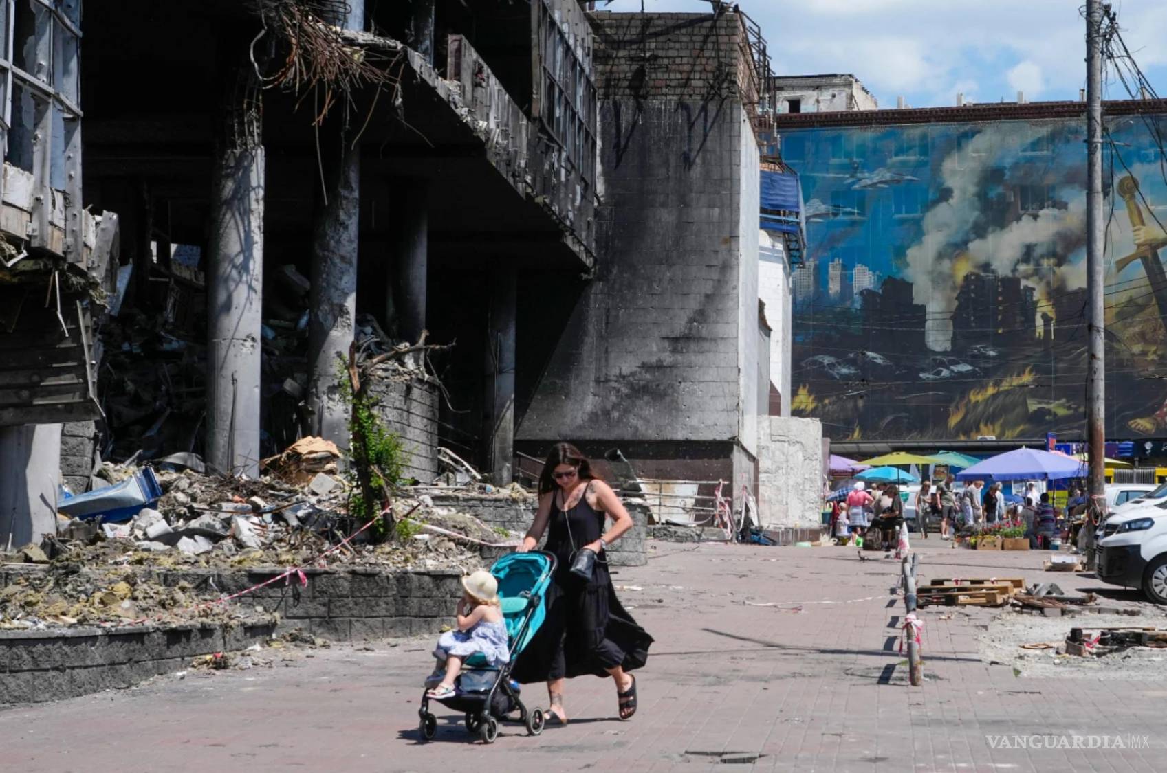 $!Una madre camina con su bebé en carriola junto a un edificio dañado cubierto de pinturas de artistas callejeros y un gran mercado urbano en Kiev, Ucrania.