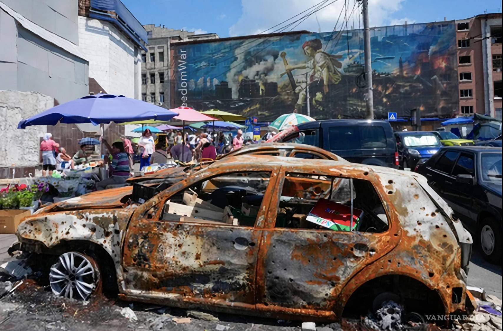 $!Gente compra comida en un mercado improvisado al aire libre, con coches quemados y fondo un mural hecho por artistas callejeros en Kiev, Ucrania.
