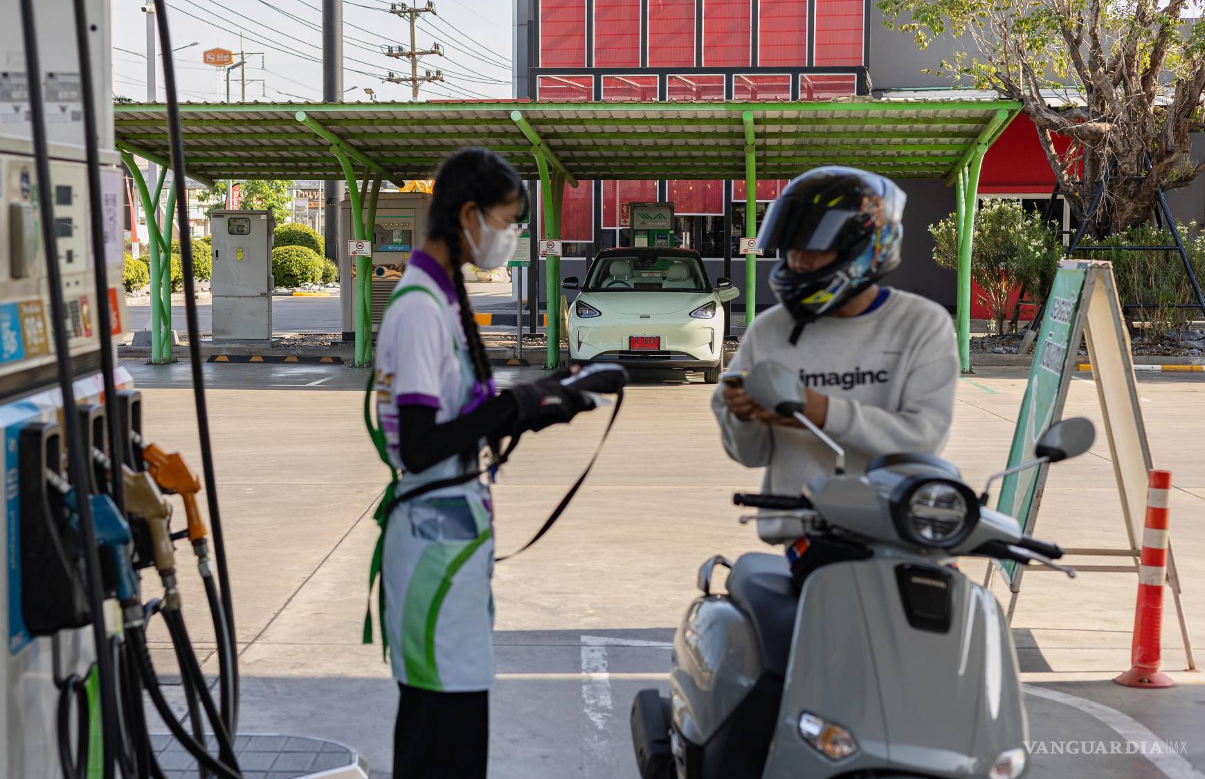 $!Un motociclista llena el tanque frente a un punto de recarga para vehículos eléctricos en una gasolinera en la provincia tailandesa de Nakorn Pathom.
