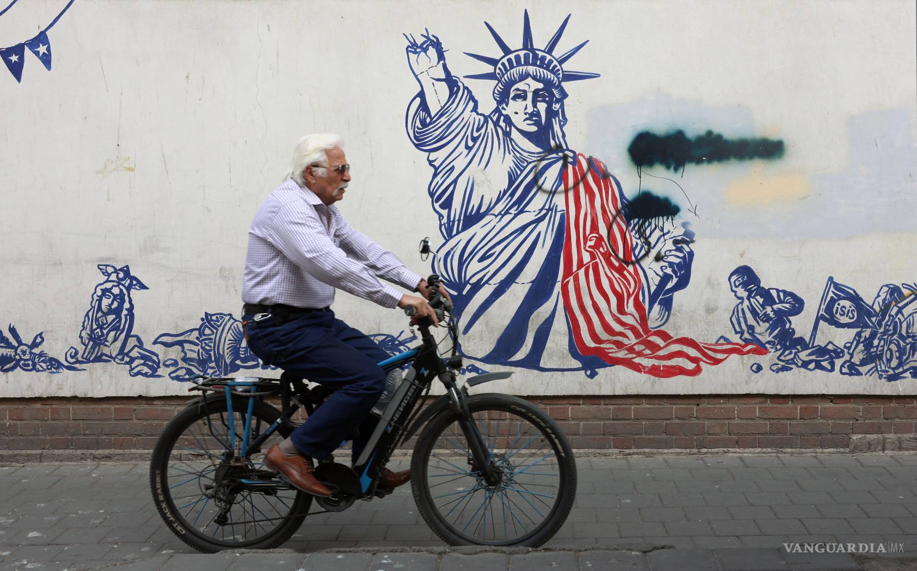 $!Un anciano iraní monta en bicicleta junto a un mural antiestadounidense en Teherán, Irán.