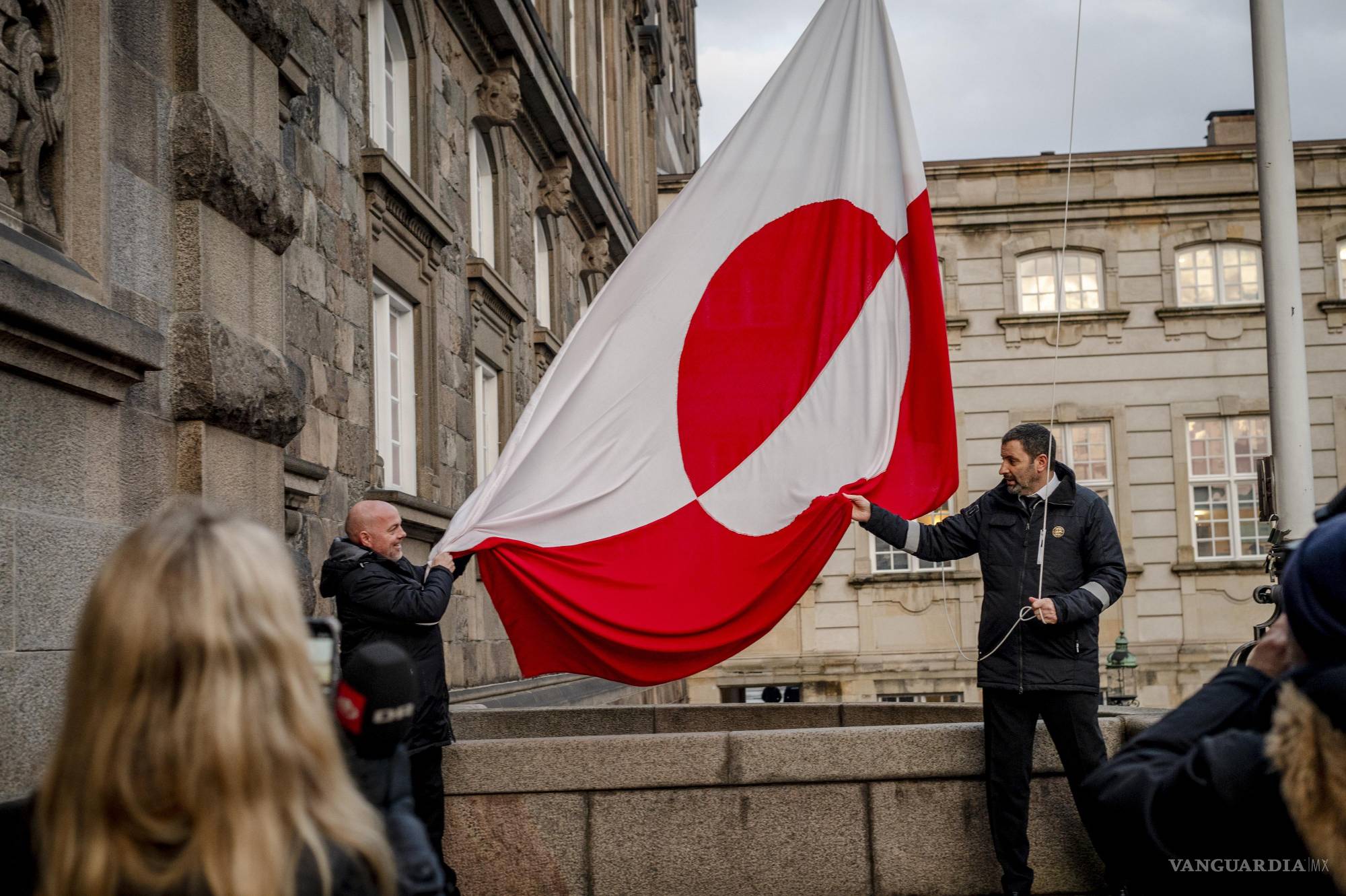 $!Dos hombres despliegan la bandera de Groenlandia frente a un edificio gubernamental, símbolo del fuerte sentimiento de soberanía que domina el debate actual.