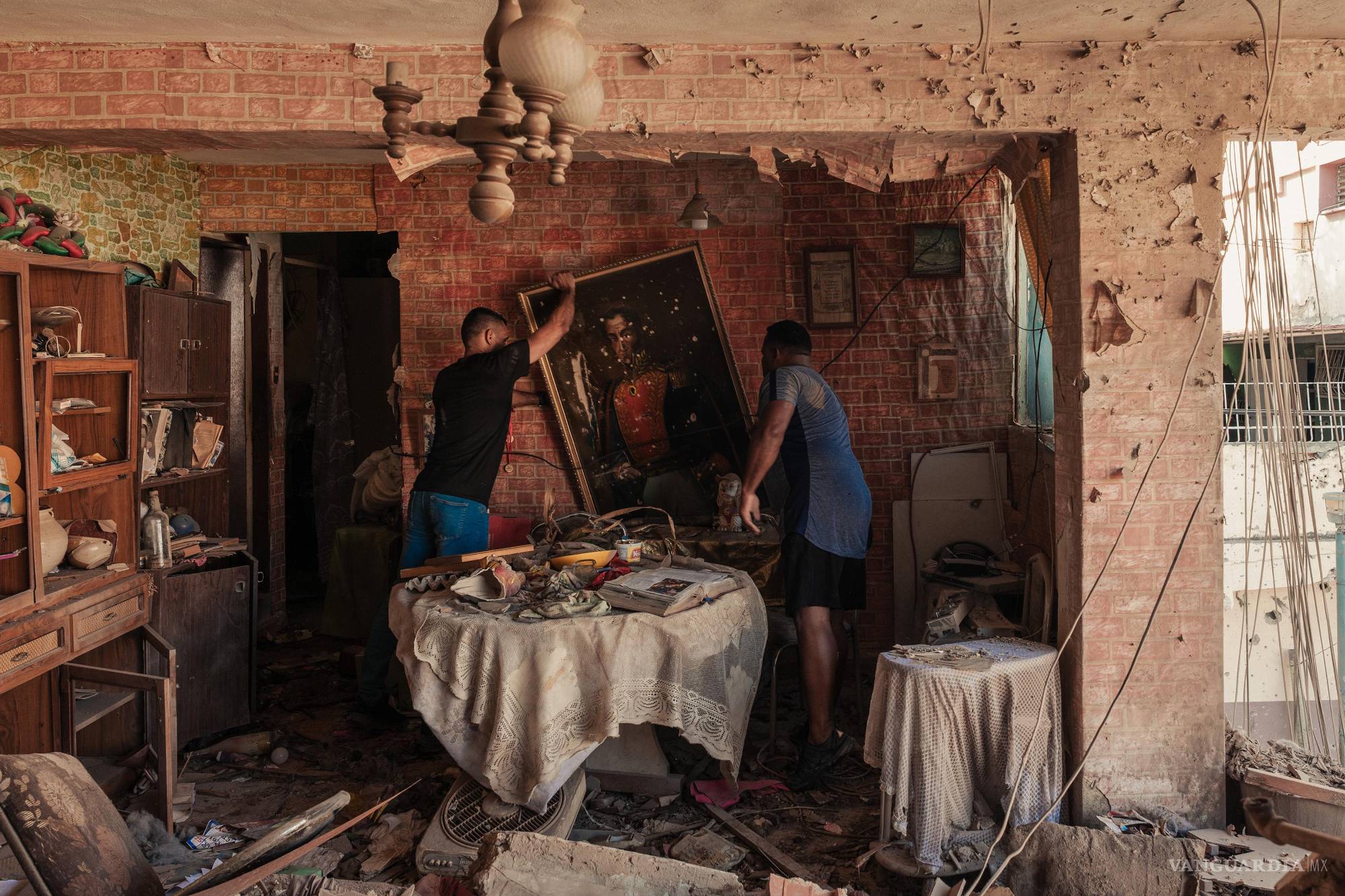 $!Dos hombres intentan rescatar pertenencias y una pintura histórica en una vivienda dañada tras los recientes acontecimientos militares en Venezuela.
