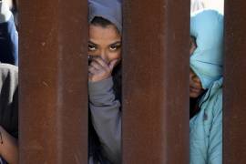 Dos migrantes que aguardan para solicitar asilo entre dos muros fronterizos miran a través de uno de ellos en San Diego.