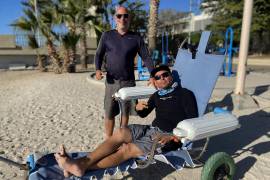 El experto en mecánica automotriz, Andrés Mata (i), y el prestador de servicios turísticos, Jairo Westle, posan junto a una silla de ruedas anfibia el 23 de abril de 2022, en una playa del balneario de La Paz (México).