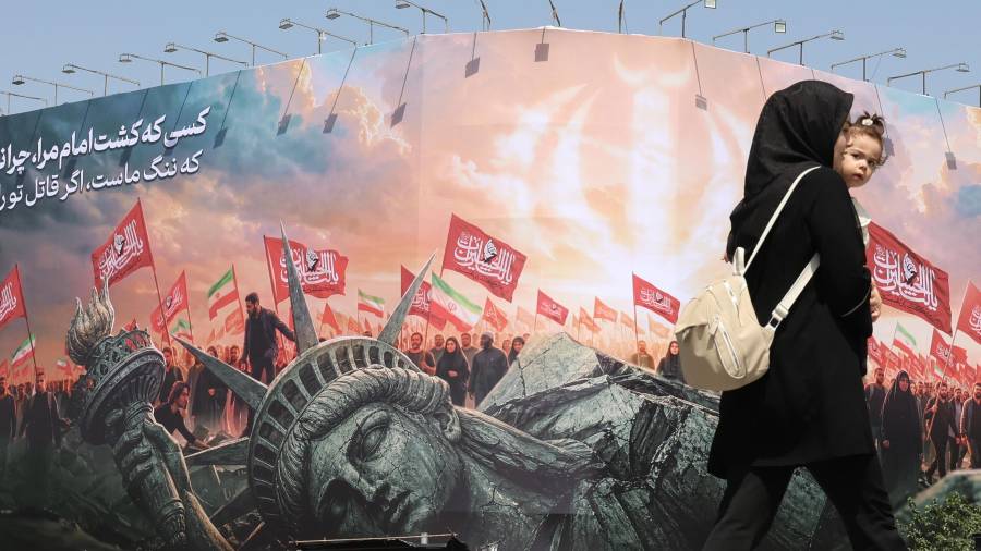 Una mujer iraní que lleva a su hijo en brazos pasa junto a una valla publicitaria antiestadounidense en la plaza Enghelab, en el centro de Teherán, Irán, el 3 de agosto de 2026.