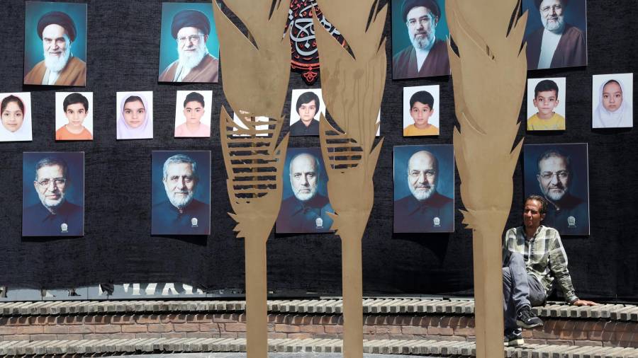 Un hombre iraní está sentado bajo un muro decorado con imágenes de líderes iraníes y de personas fallecidas en la guerra, incluidos niños de Minab, en las calles de Teherán, la capital de Irán.
