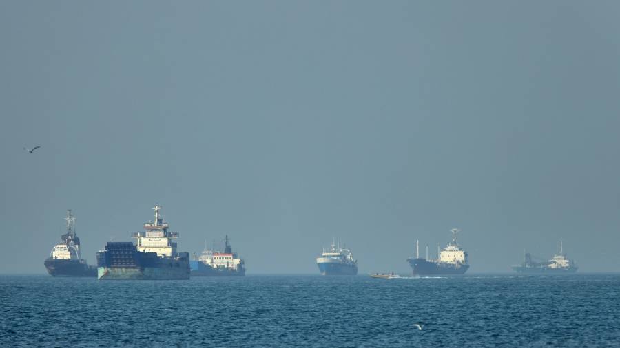 Buques de carga, incluyendo graneleros y buques de carga general, permanecen anclados en alta mar mientras una pequeña lancha motora pasa en primer plano, en el estrecho de Ormuz.