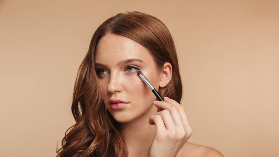 Beauty portrait of mystery smiling ginger woman with long hair looking away while applying cosmetics with brush for eyeshadow over cream background