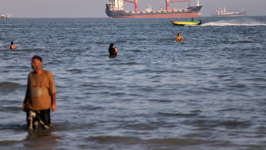 Residentes nadan en las aguas del estrecho de Ormuz mientras una pequeña lancha pasa junto a cargueros y otros buques comerciales en alta mar, cerca de Bandar Abbas, Irán.