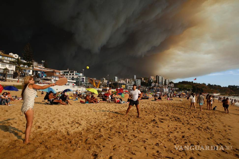 $!Turistas juegan a las palas en una playa con una gran nube de humo negro a sus espaldas, procedente de los incendios forestales, en Viña del Mar, Chile.