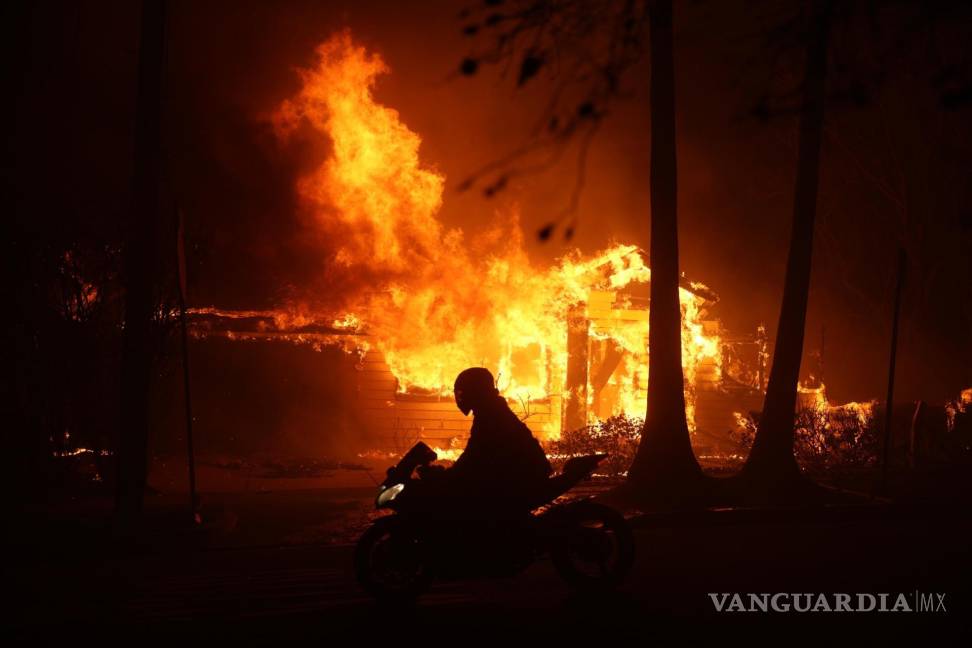 $!Una persona conduce su motocicleta junto a una casa en llamas durante los incendios en Palisades , California, Estados Unidos.