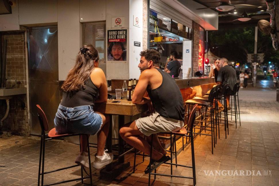 $!Dos personas charlan en un bar junto a un cartele que pide la liberación de los rehenes retenidos por Hamás en Gaza desde hace casi un año, en Tel Aviv, Israel.