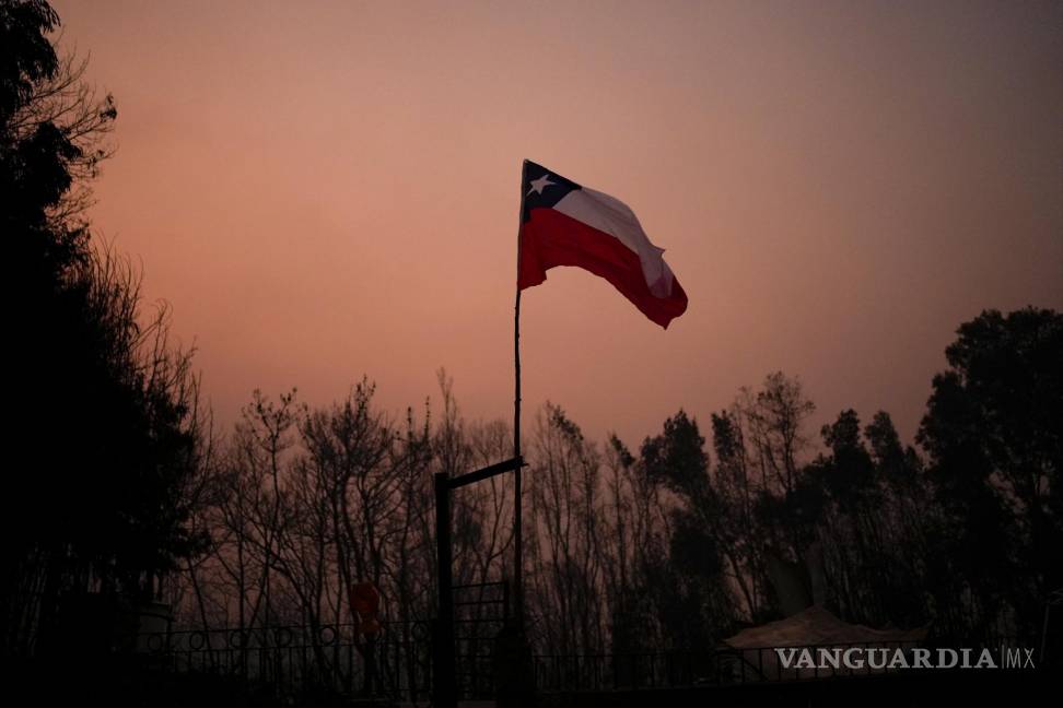 $!Una bandera chilena colocada en medio de árboles carbonizados bajo un cielo rojo causado por incendios forestales en Santa Juana, Chile.