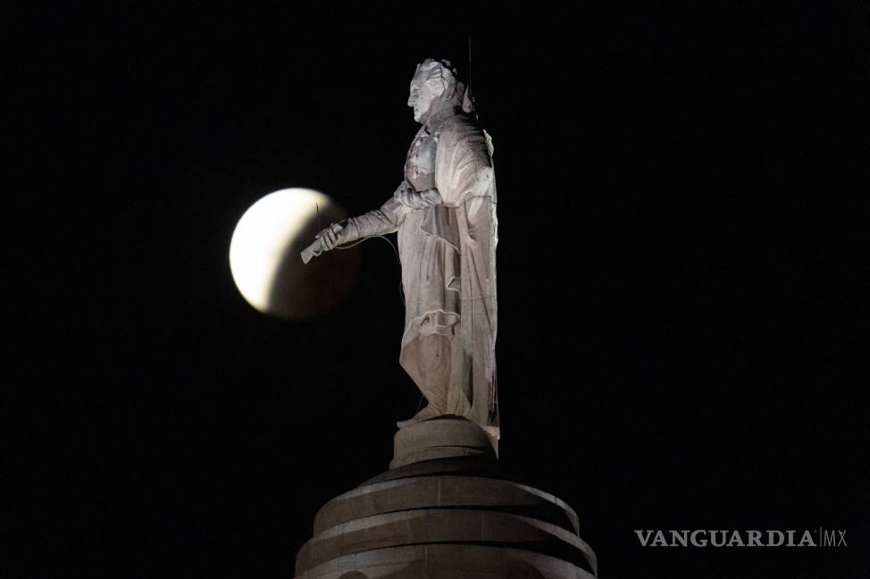 $!La sombra de la tierra cubre la luna llena durante un eclipse lunar parcial visible cerca de una estatua de George Washington en lo alto del Monumento a Washington de Baltimore en Baltimore. AP/Julio Cortez